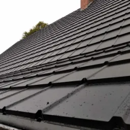 Nowoczesne, czarne pokrycie dachowe z blachy na budynku z kominem z cegły w tle, widoczne krople wody po deszczu.