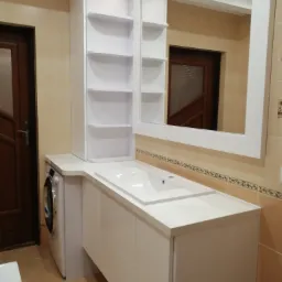 Biała umywalka z szafką i lustrem w łazience, obok pralka i wysoki regał z półkami, brązowe płytki na ścianach.