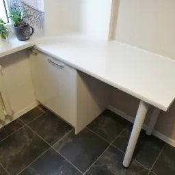 Biały blat z szafką, zamontowany przy oknie z roślinami doniczkowymi i grzejnikiem, na podłodze ciemne płytki.