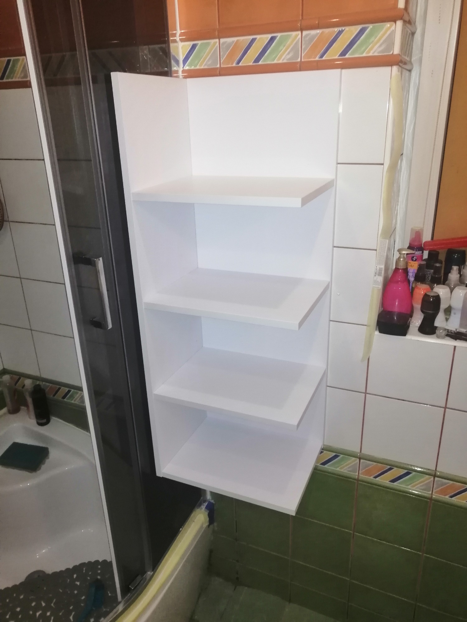 Biała, narożna półka łazienkowa z trzema poziomami, zamontowana na ścianie obok kabiny prysznicowej, z widocznymi kosmetykami na półce i kafelkami w tle.