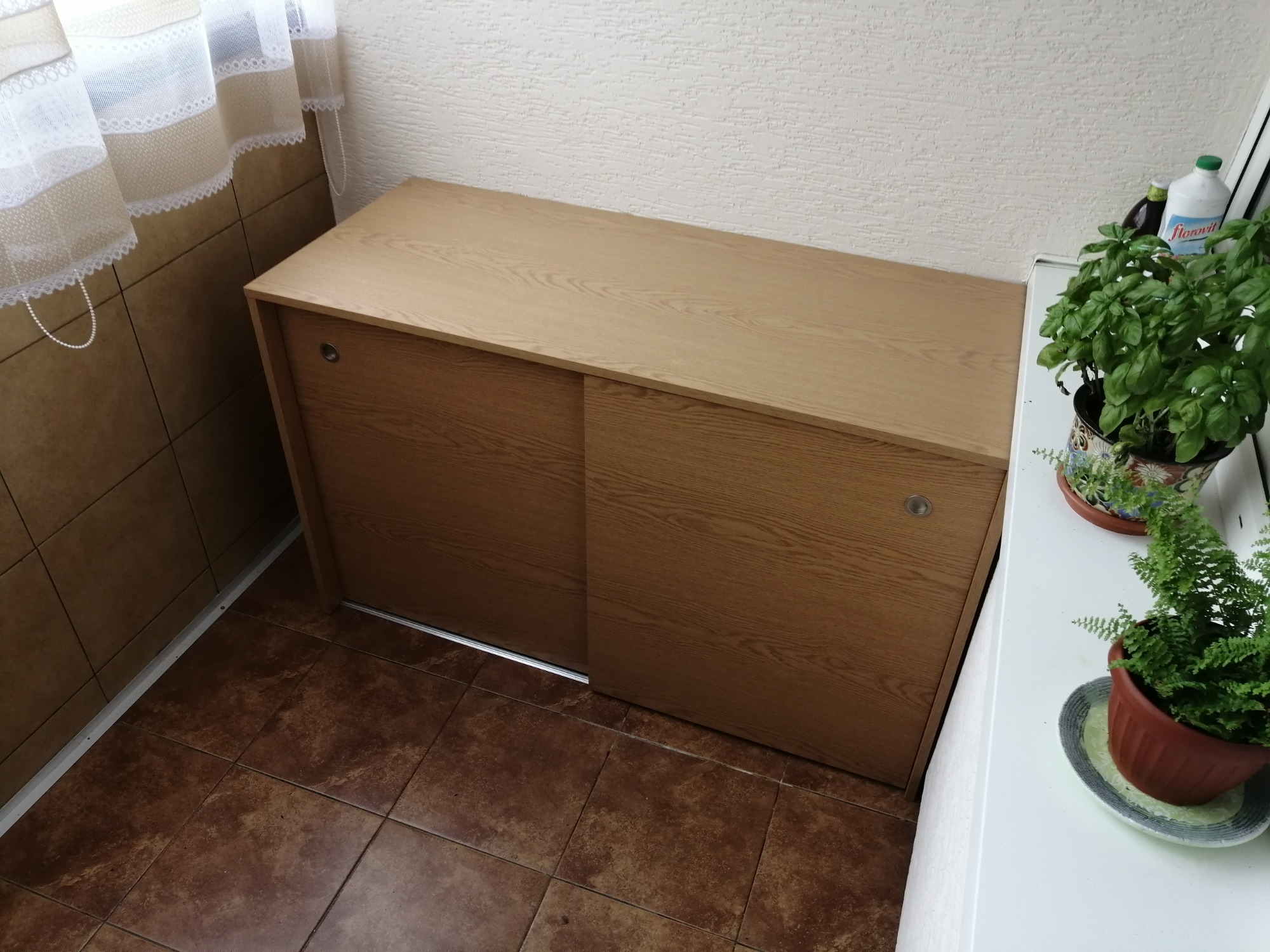 Szafka z przesuwnymi drzwiami z jasnego drewna na balkonie z brązowymi płytkami, obok rośliny w doniczce i zasłony.