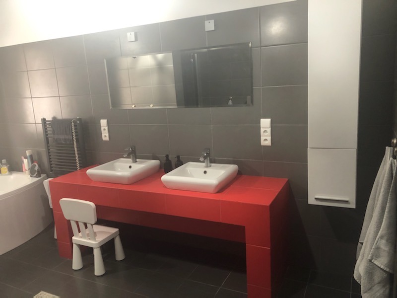 Nowoczesna łazienka z dwoma umywalkami na czerwonym blacie, szare płytki ścienne, biała szafka wisząca i małe białe krzesełko.