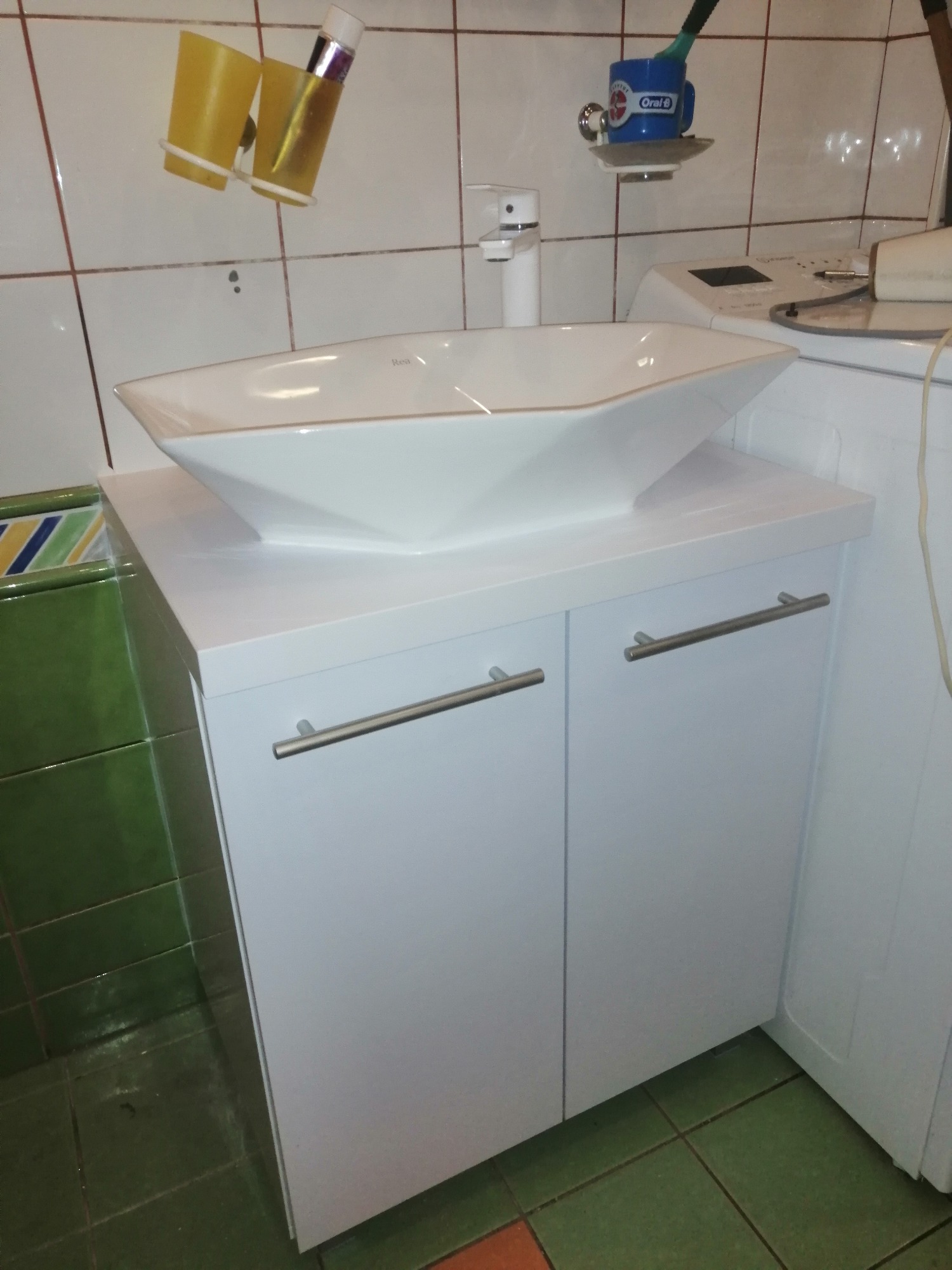 Biała szafka łazienkowa z umywalką w kształcie łodzi, srebrne uchwyty, obok pralka, widoczne akcesoria łazienkowe na ścianie z brązowymi kaflami.