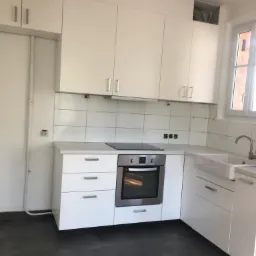 Jasna kuchnia z białymi szafkami, płytkami i szarą podłogą, widoczne podłączenia elektryczne i hydrauliczne przed wykończeniem.