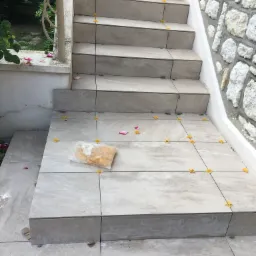 Schody zewnętrzne wyłożone jasnymi płytkami ceramicznymi, z użyciem żółtych krzyżyków dystansowych i paczki z zaprawą klejową na podeście, obok ściana z kamienia naturalnego.