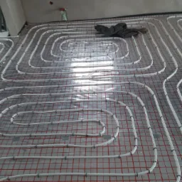 Białe rury ogrzewania podłogowego ułożone w pętle na czerwonej siatce, widoczne mocowania i fragment ubrania w rogu pomieszczenia, surowy stan wykończenia.