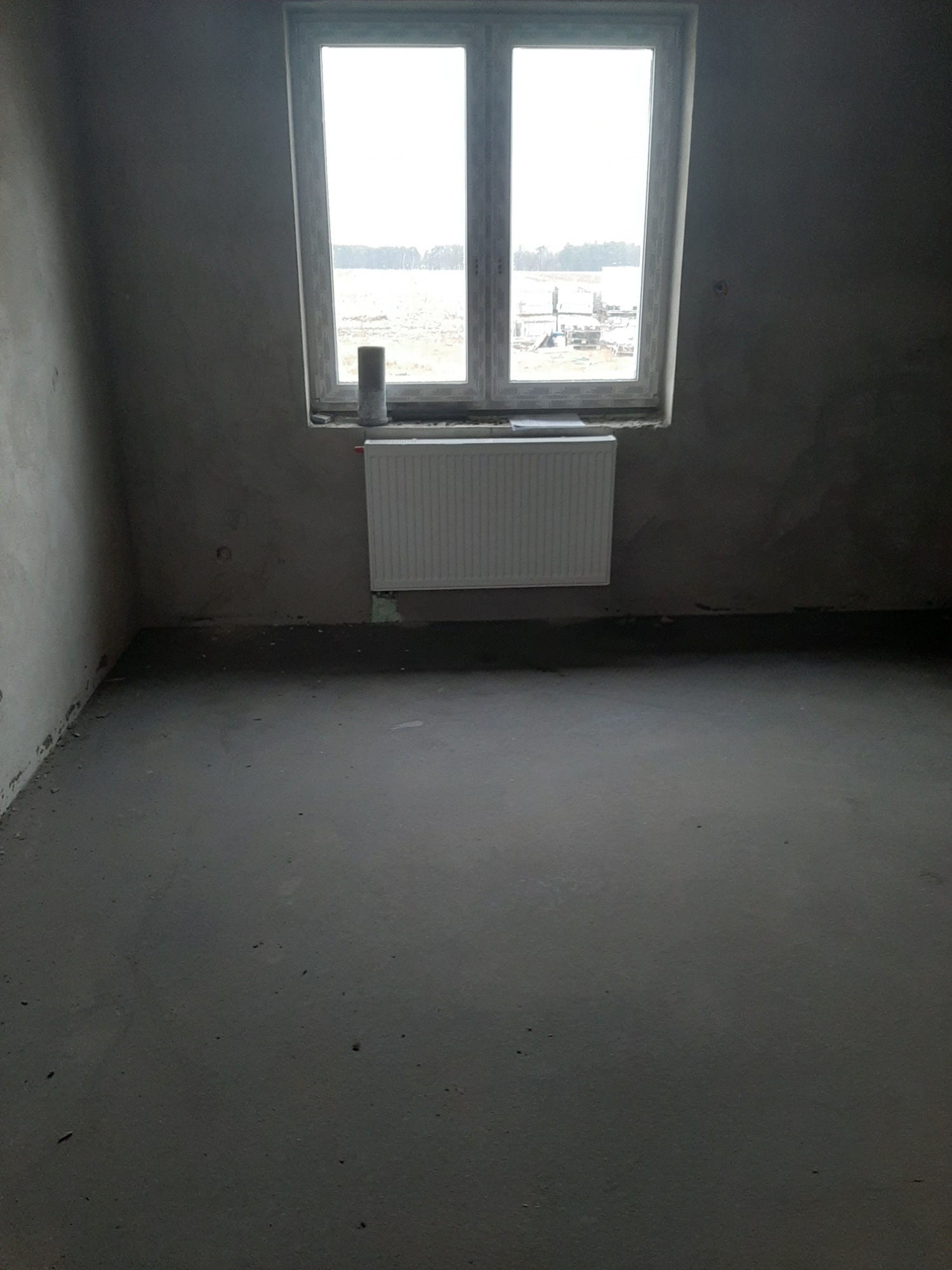 Surowe wnętrze pokoju w trakcie remontu, z grzejnikiem pod oknem z widokiem na krajobraz za oknem.