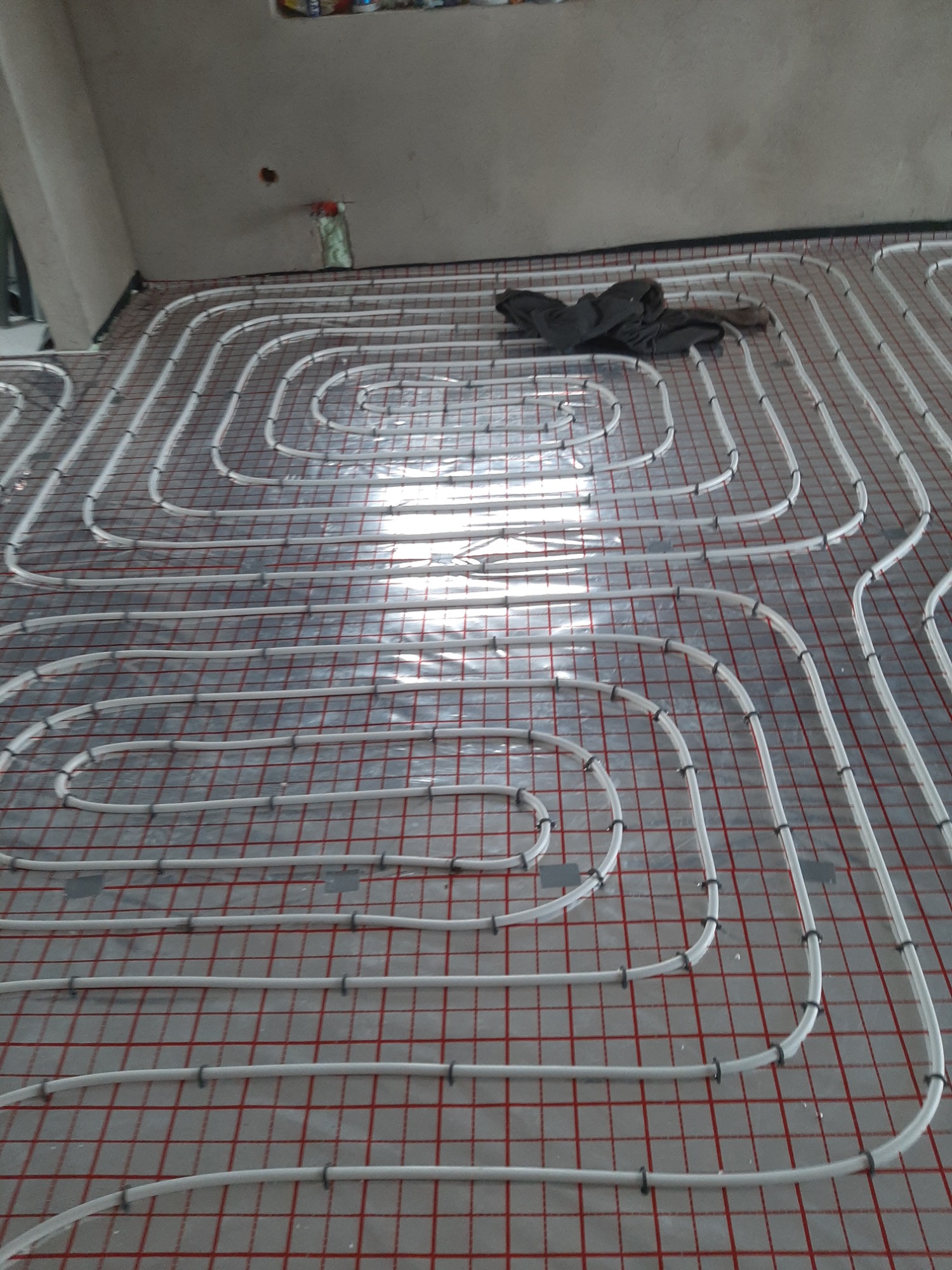 Białe rury ogrzewania podłogowego ułożone w pętle na czerwonej siatce, widoczne mocowania i fragment ubrania w rogu pomieszczenia, surowy stan wykończenia.