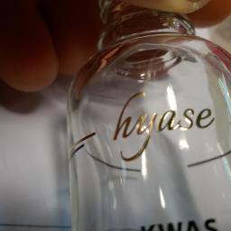 GRAF-PAK - Szklana butelka z złotym napisem 'hyase' i czarnymi, cienkimi liniami dekoracyjnymi, uchwycona z bliska pod kątem, z widocznym fragmentem dłoni w tle.