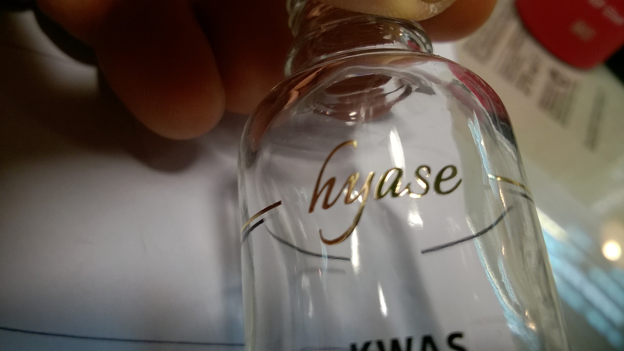Szklana butelka z złotym napisem 'hyase' i czarnymi, cienkimi liniami dekoracyjnymi, uchwycona z bliska pod kątem, z widocznym fragmentem dłoni w tle.