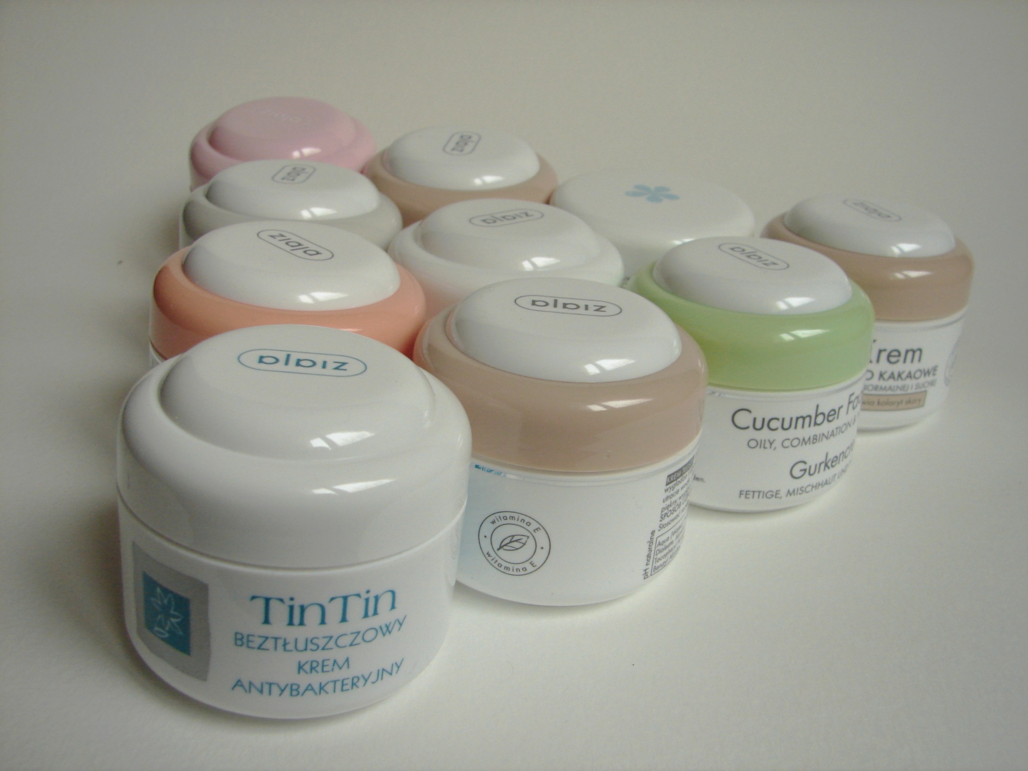 Zestaw kremów do twarzy różnych marek, w okrągłych, białych opakowaniach z kolorowymi pokrywkami (różowa, beżowa, zielona), na białym tle.