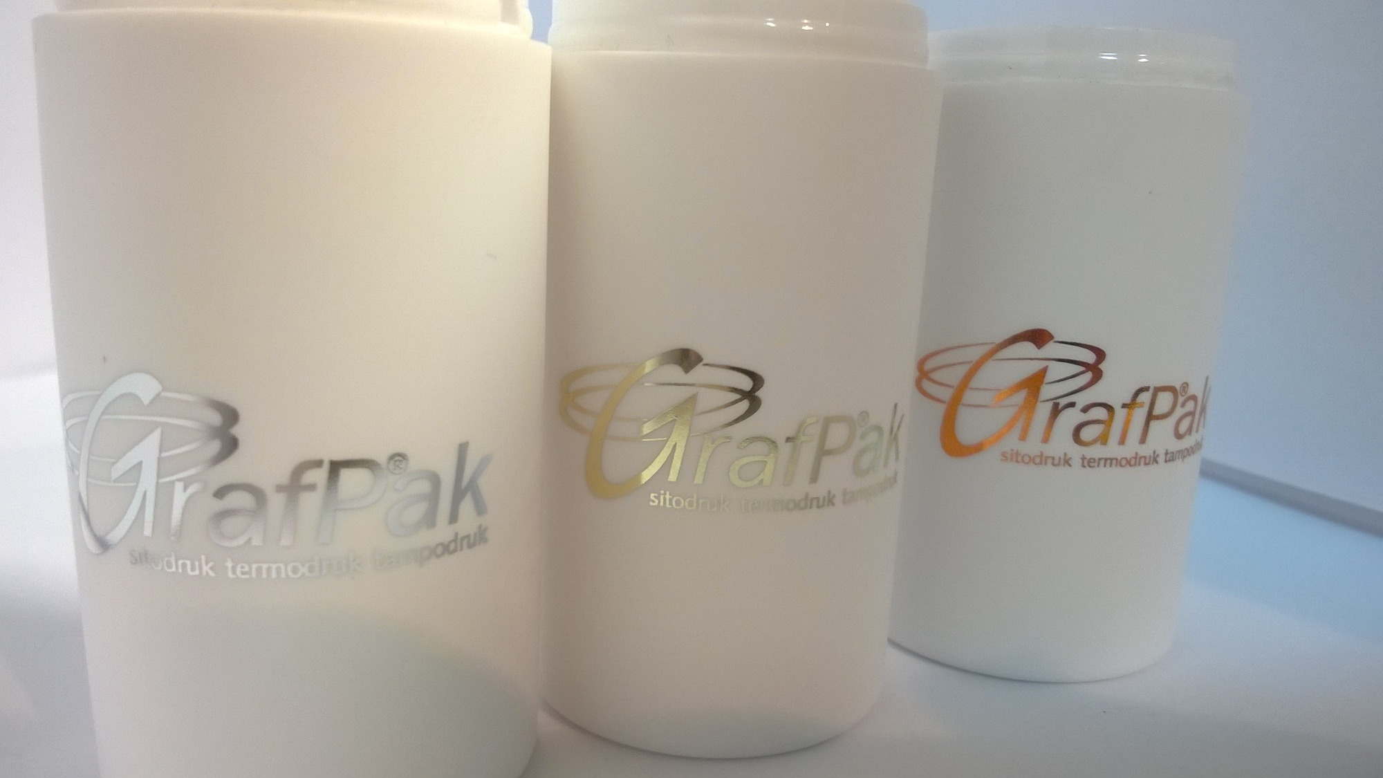 Trzy białe pojemniki z nadrukowanym logo firmy GrafPak w kolorach srebrnym, złotym i miedzianym, wykonanym techniką sitodruku, termodruku i tampodruku.