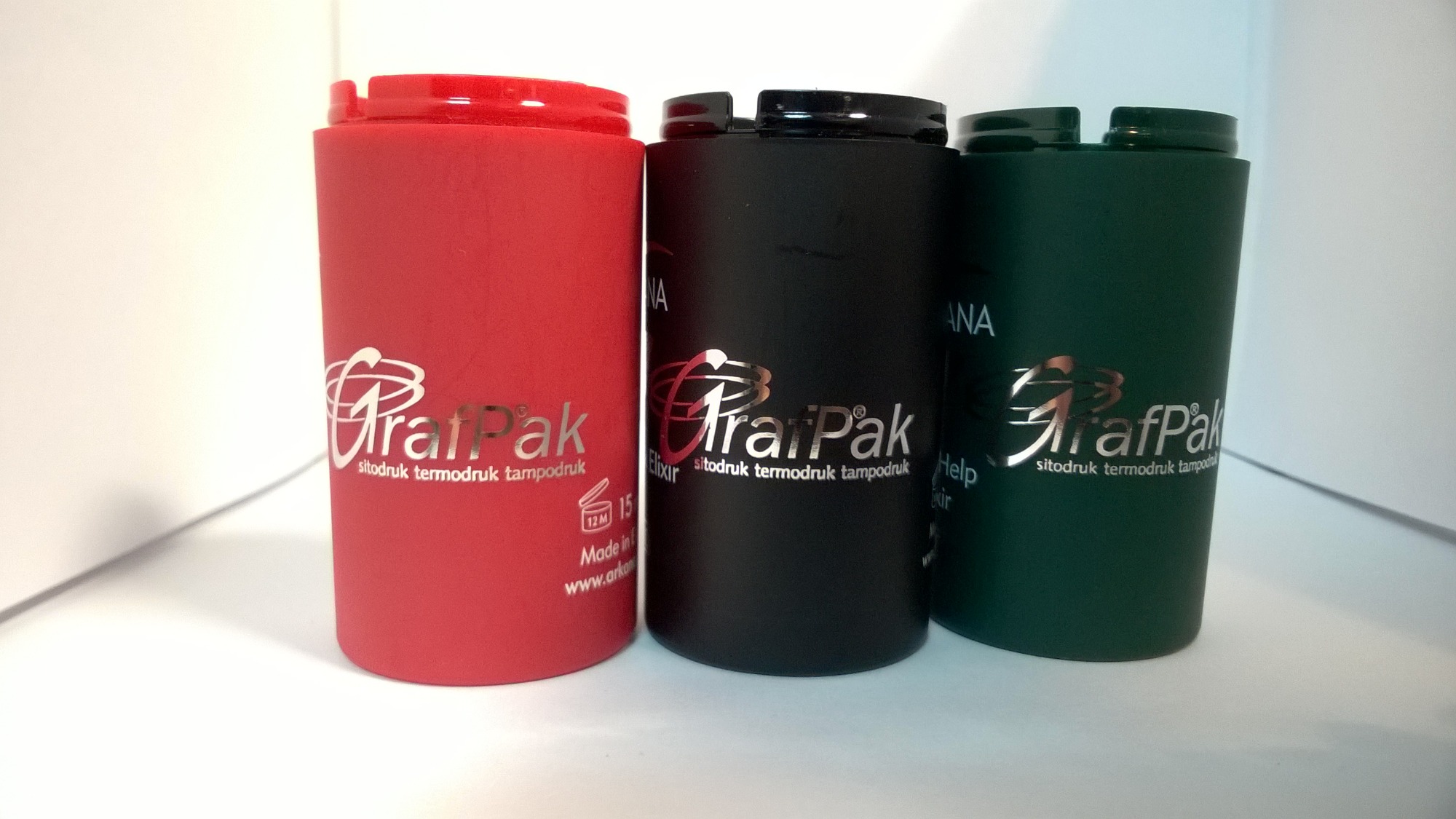 Trzy cylindryczne pojemniki w kolorach czerwonym, czarnym i zielonym z nadrukowanym srebrnym logo 'GrafPak sitodruk termodruk tampodruk' na białym tle.