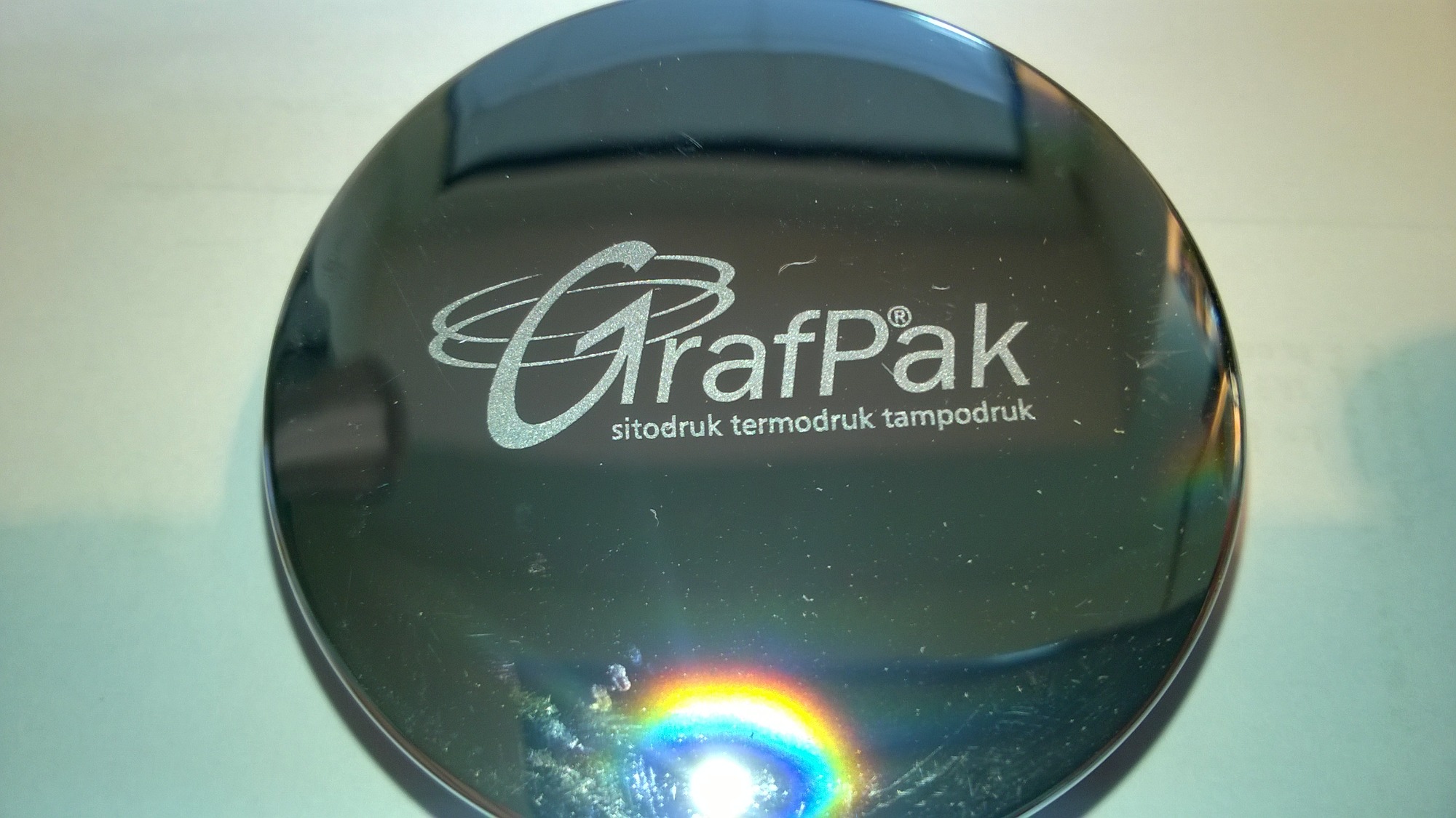 Błyszcząca, okrągła powierzchnia z nadrukiem logo 'GrafPak' i informacją o metodach druku: sitodruk, termodruk, tampodruk, z widocznym odbiciem tęczy na dole.