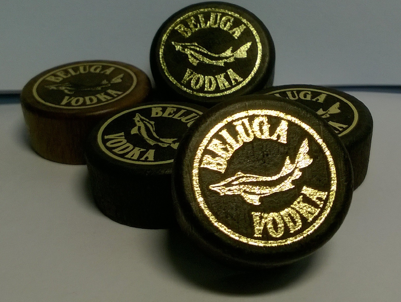 Zbliżenie na grupę drewnianych kapsli z nadrukiem logo Beluga Vodka, z wyraźnie widocznym złotym rysunkiem bieługi i napisem, na jasnym tle.
