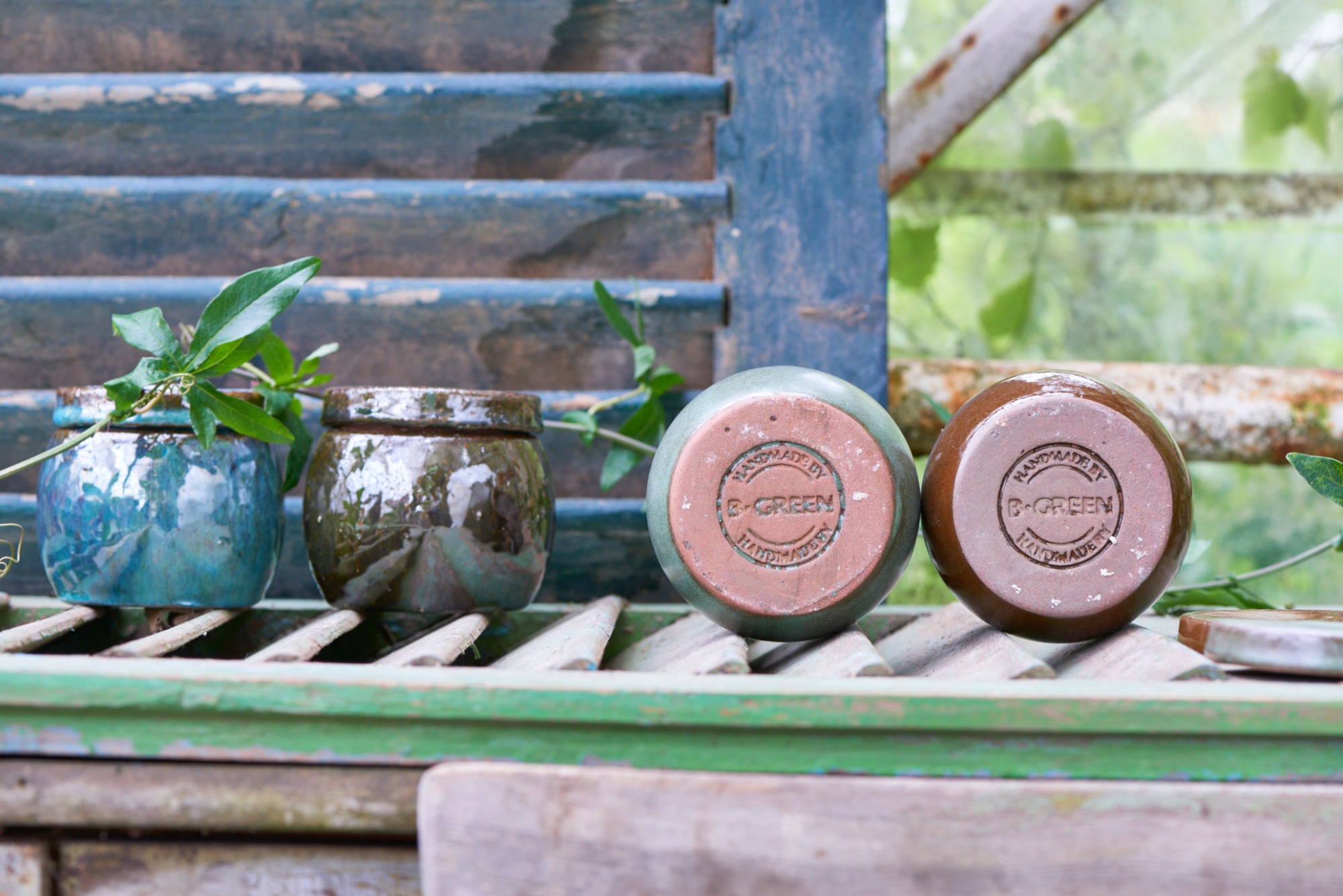 Ręcznie robione, szkliwione doniczki ceramiczne w odcieniach błękitu i brązu, z widocznym stemplem producenta 'B-GREEN' na spodzie, ustawione na drewnianej, pomalowanej na zielono konstrukcji...