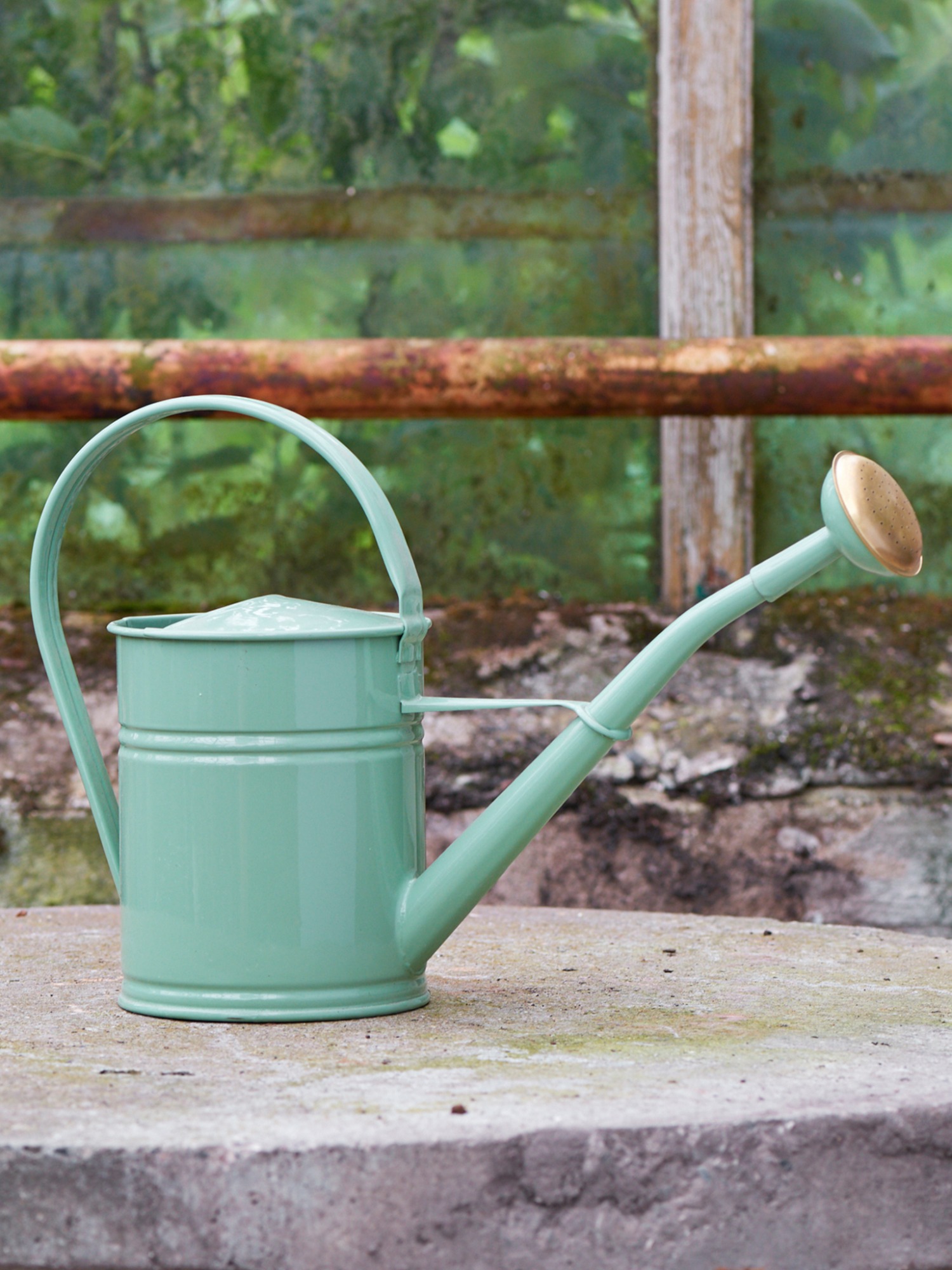 Pastelowa, miętowa konewka z mosiężną końcówką na betonowym blacie, na tle starego okna szklarni porośniętego mchem.