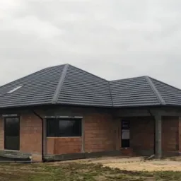 Nowo wybudowany dom jednorodzinny z ciemną dachówką, widoczne rynny i okno dachowe, ściany w trakcie budowy z czerwonej cegły, pochmurne niebo w tle.