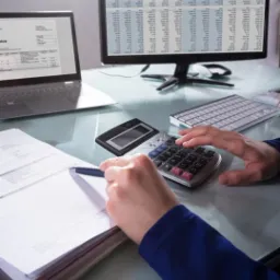 Kobieta w Płocku oblicza dane na kalkulatorze, mając przed sobą stertę dokumentów, laptopa i monitor z arkuszem kalkulacyjnym.