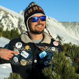 Buttonfly - Uśmiechnięty mężczyzna w czapce i okularach przeciwsłonecznych, ubrany w sweter ozdobiony przypinkami, wskazujący palcem na jedną z nich, na tle ośnieżonych gór i zielonych drzew iglastych.