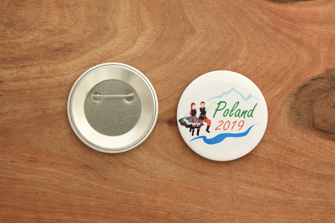 Dwa okrągłe znaczki na drewnianym tle: jeden z grafiką pary w strojach ludowych, napisem 'Poland 2019' i stylizowanymi górami, drugi z widocznym zapięciem agrafkowym.