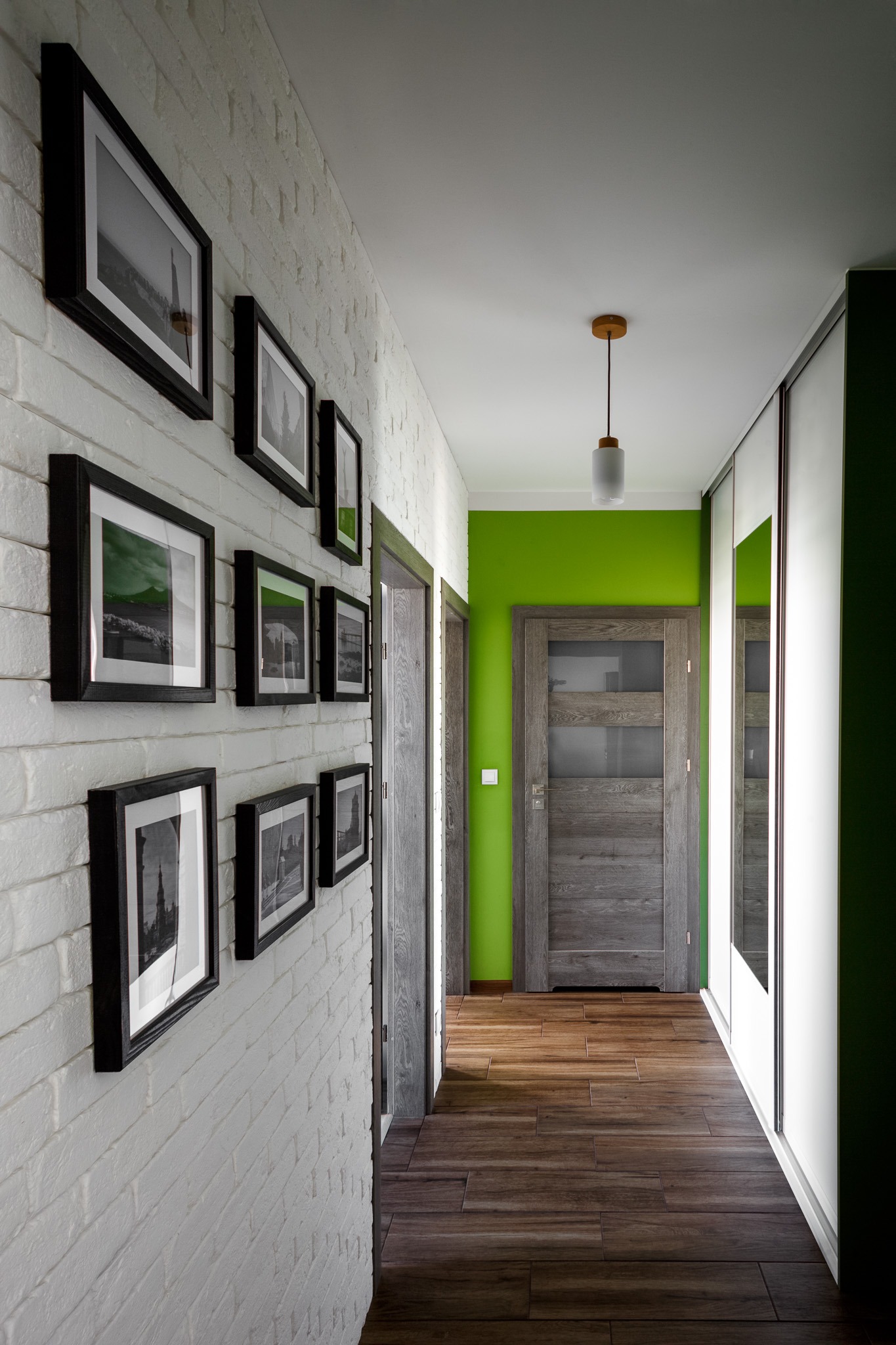 Nowoczesny korytarz z białą cegłą na ścianie, drewnianą podłogą, zieloną ścianą w tle i galerią czarno-białych zdjęć w ramkach.