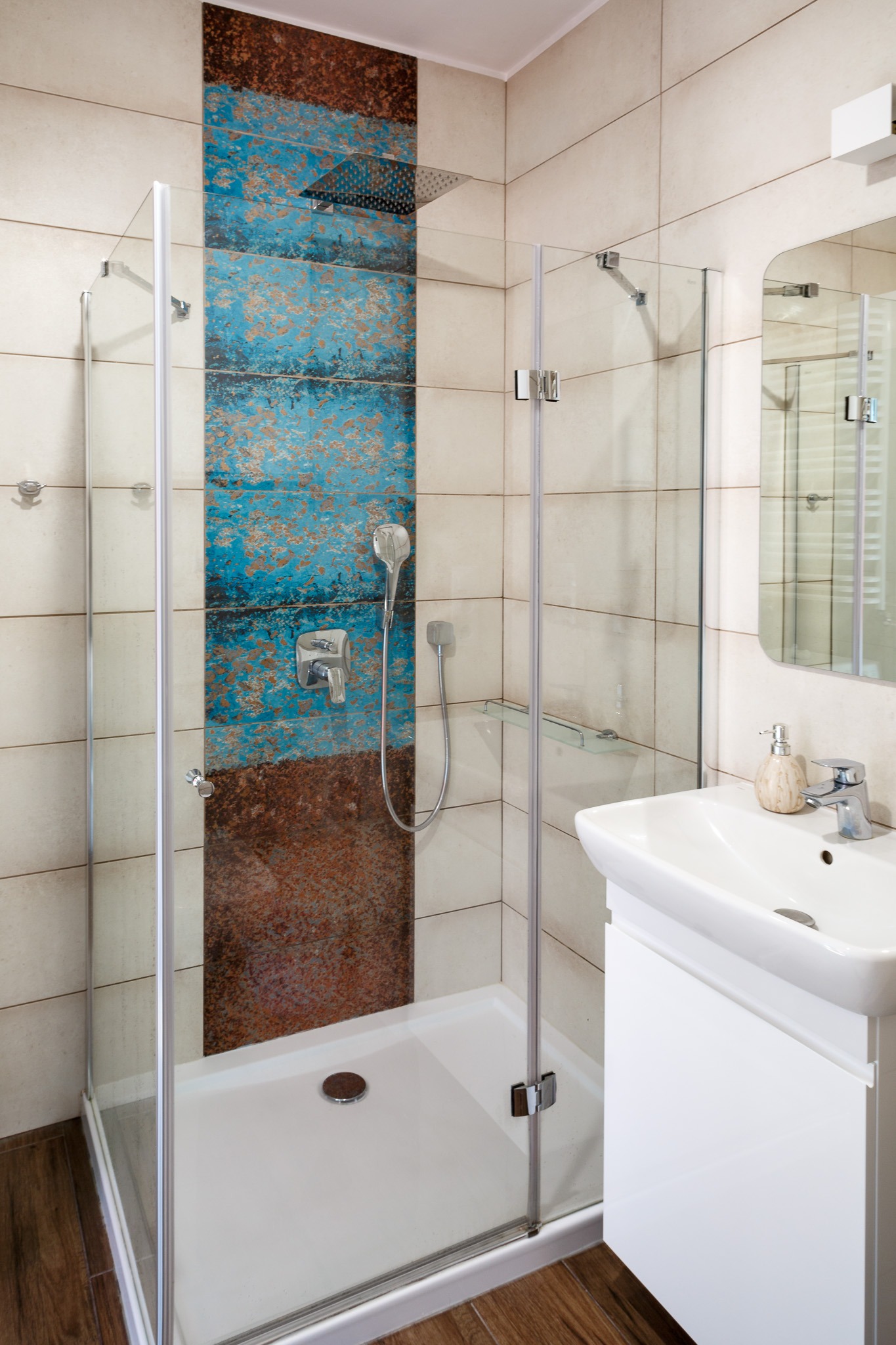 Nowoczesna kabina prysznicowa ze szklanymi drzwiami i panelami z płytek imitujących rdzę i turkus, biała umywalka z szafką i lustro.