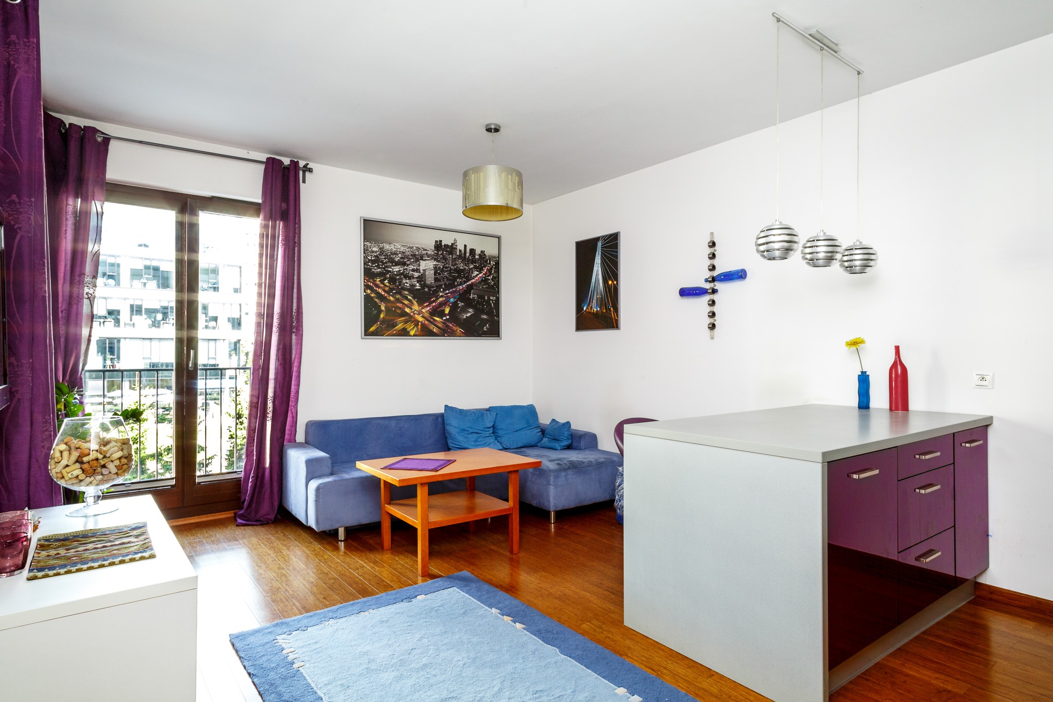 Jasne wnętrze mieszkania z niebieską sofą, drewnianym parkietem i fioletowymi akcentami, w tym zasłonami i elementami zabudowy kuchennej.
