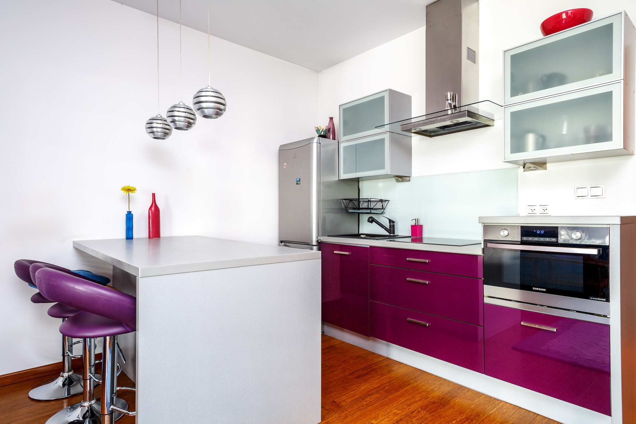 Nowoczesna kuchnia z fioletowymi szafkami, drewnianą podłogą i srebrnymi dodatkami, oświetlona trzema kulistymi lampami wiszącymi.