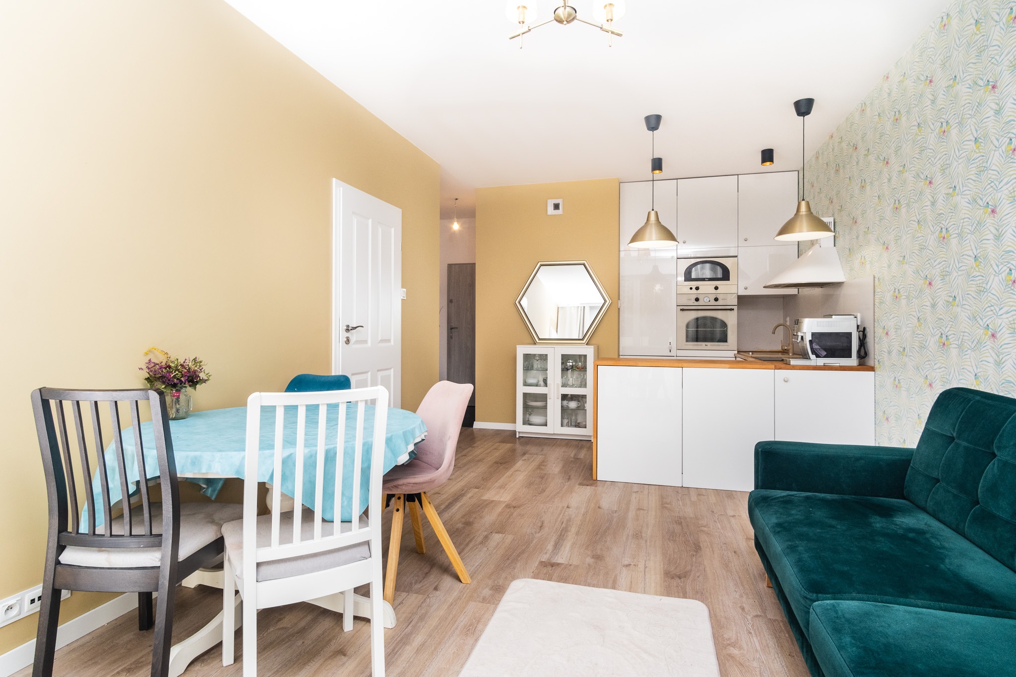 Jasne wnętrze mieszkania z aneksem kuchennym, stołem z kwiatami i zieloną sofą. Na ścianie żółta farba i tapeta w tropikalne liście.