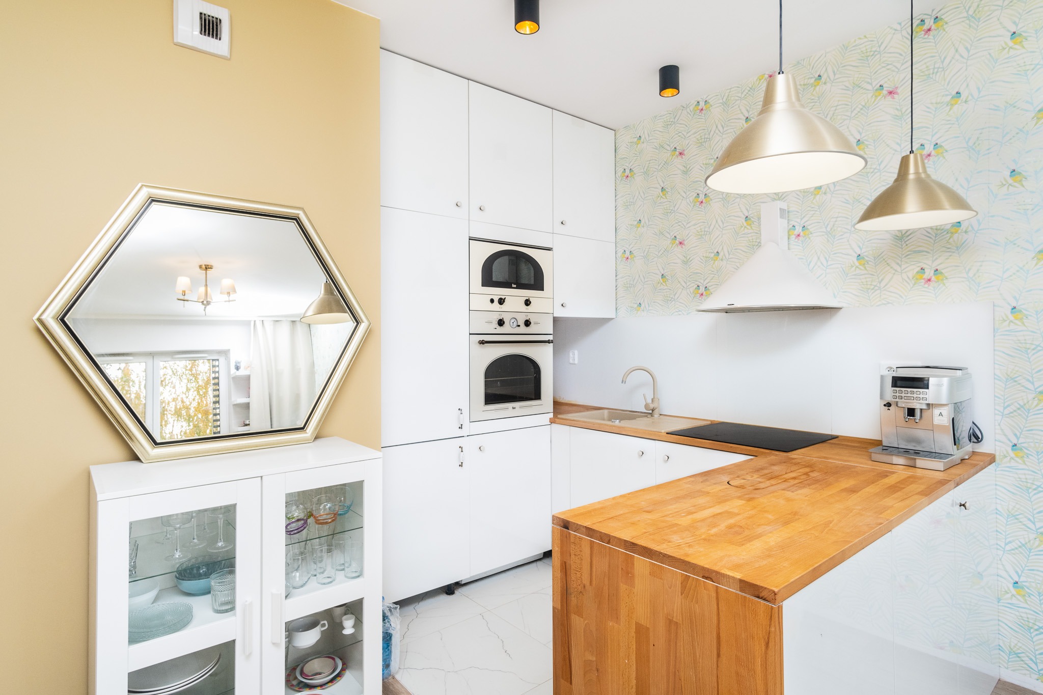 Jasna kuchnia z białymi szafkami, drewnianym blatem i lustrem w heksagonalnej ramie odbijającym żyrandol.