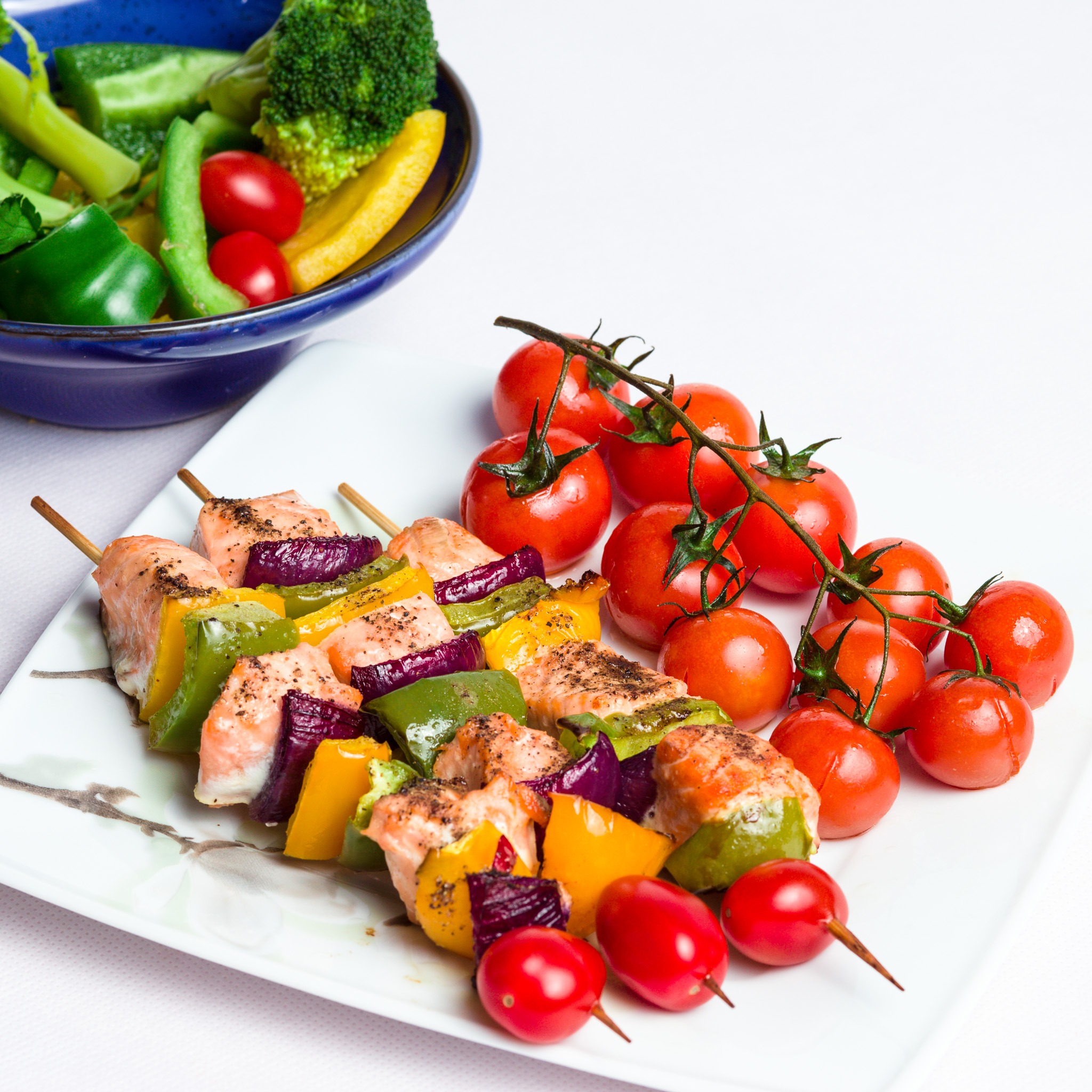 Grillowane szaszłyki z łososia i warzyw (papryka, cebula) na białym talerzu z motywem kwiatowym, obok gałązka pomidorów koktajlowych i miseczka z zielonymi warzywami.