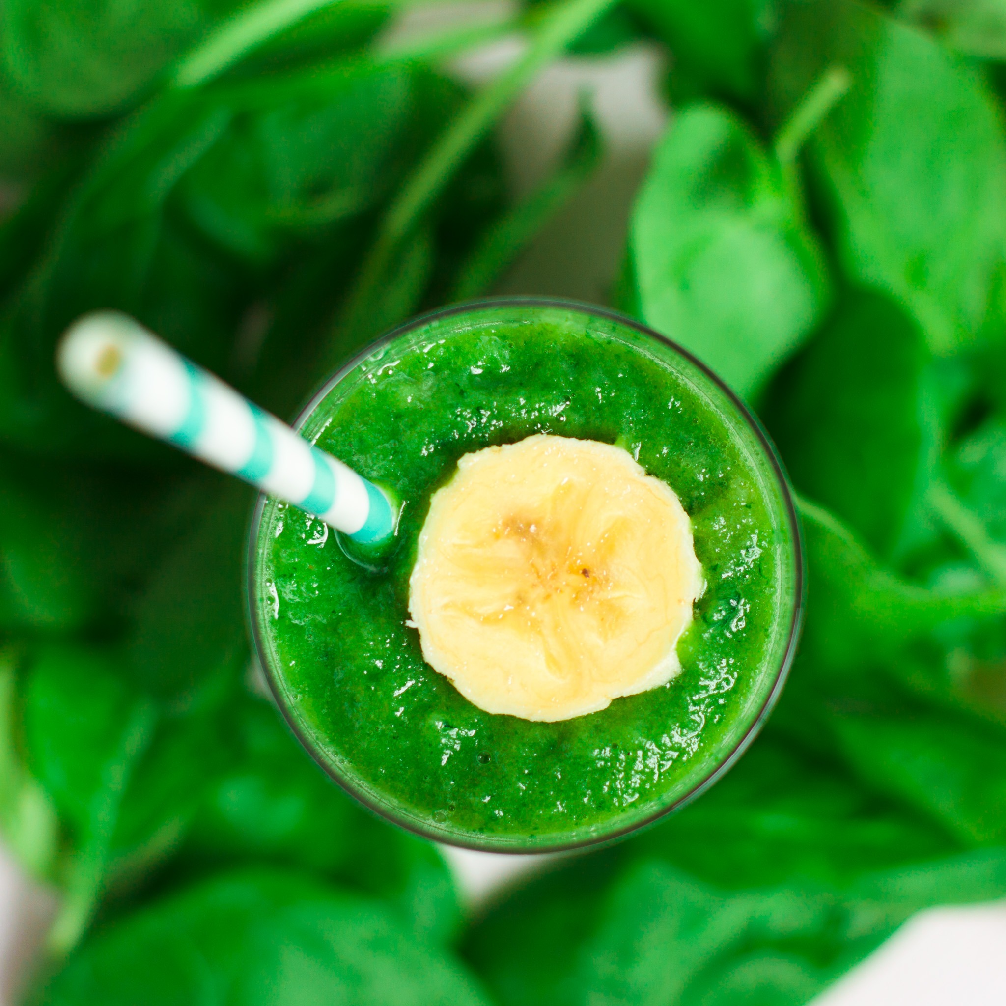 Zielony koktajl ze szpinaku i banana, ozdobiony plasterkiem banana, z papierową słomką w paski, na tle świeżych liści szpinaku, widok z góry.