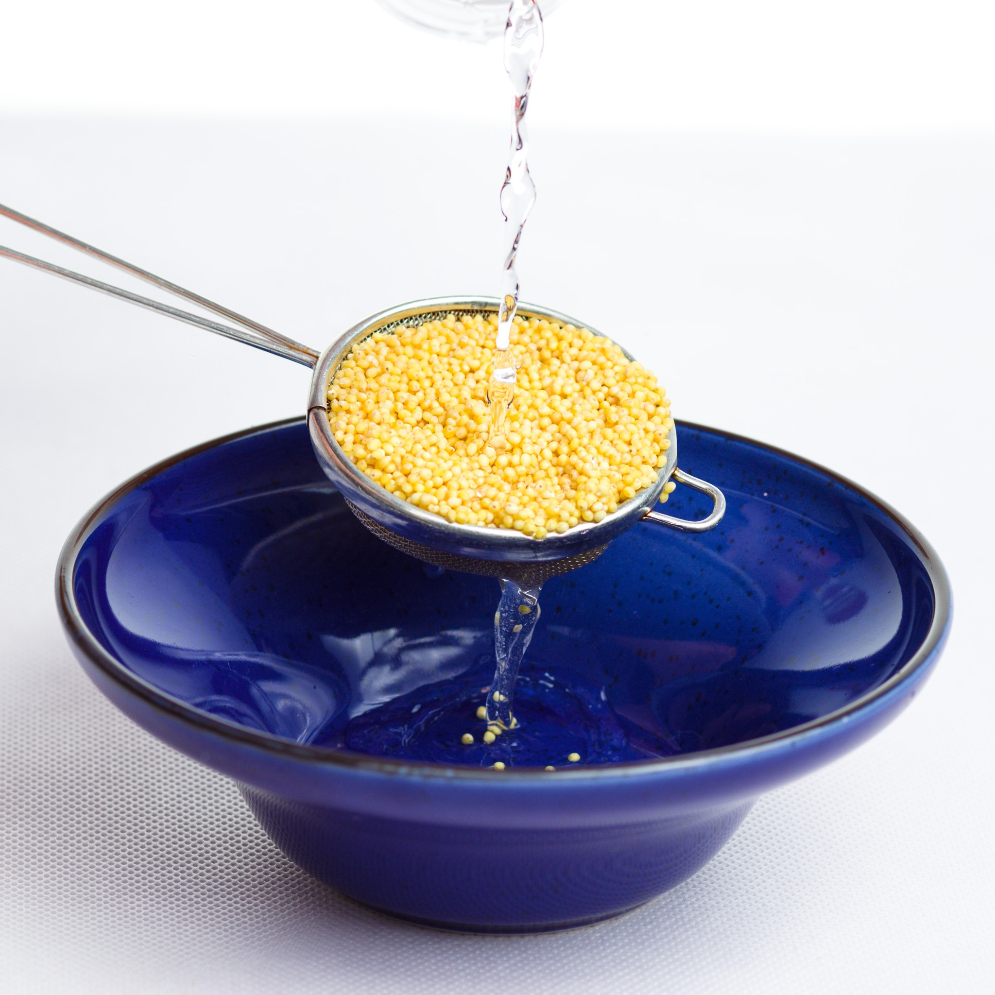 Przemywanie kaszy jaglanej w metalowym sitku nad głębokim, niebieskim talerzem, z którego widać spływającą wodę.