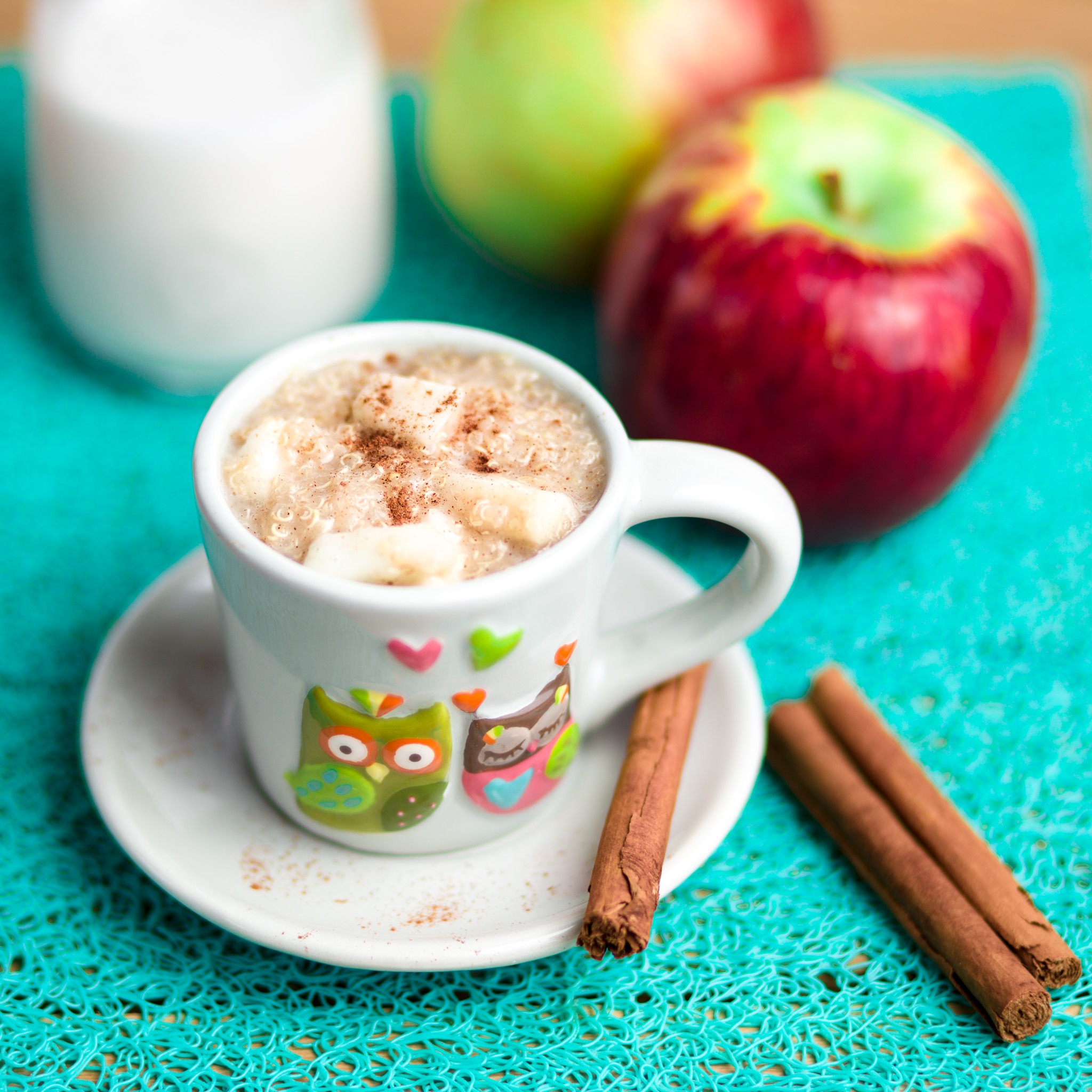 Owsianka z bananem i cynamonem w kubku z sowami na turkusowej podkładce, jabłka i mleko w tle.