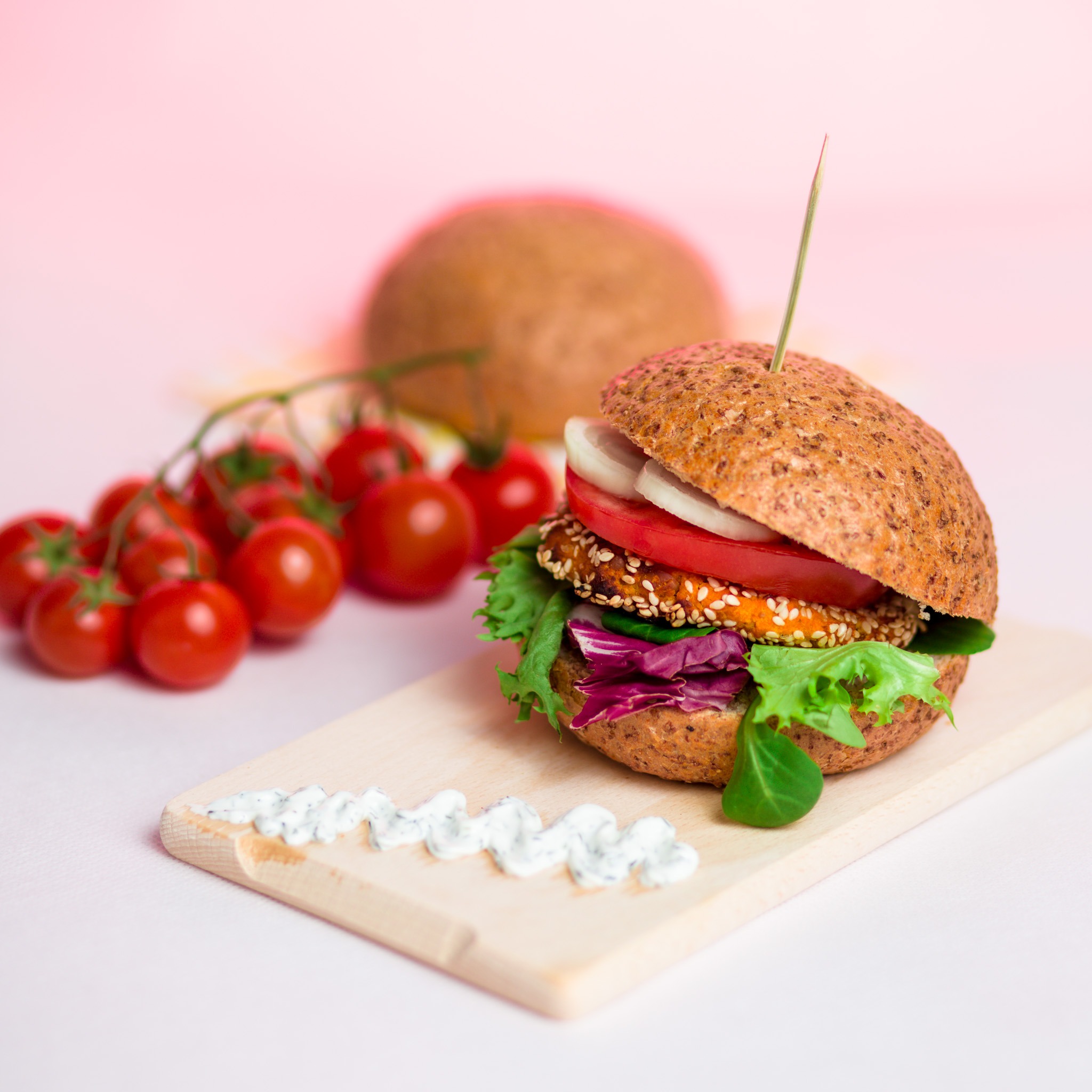 Burger z kotletem w sezamie, pomidorem, cebulą i sałatą, podany na drewnianej desce z sosem, obok pomidorki cherry i bułka w tle.