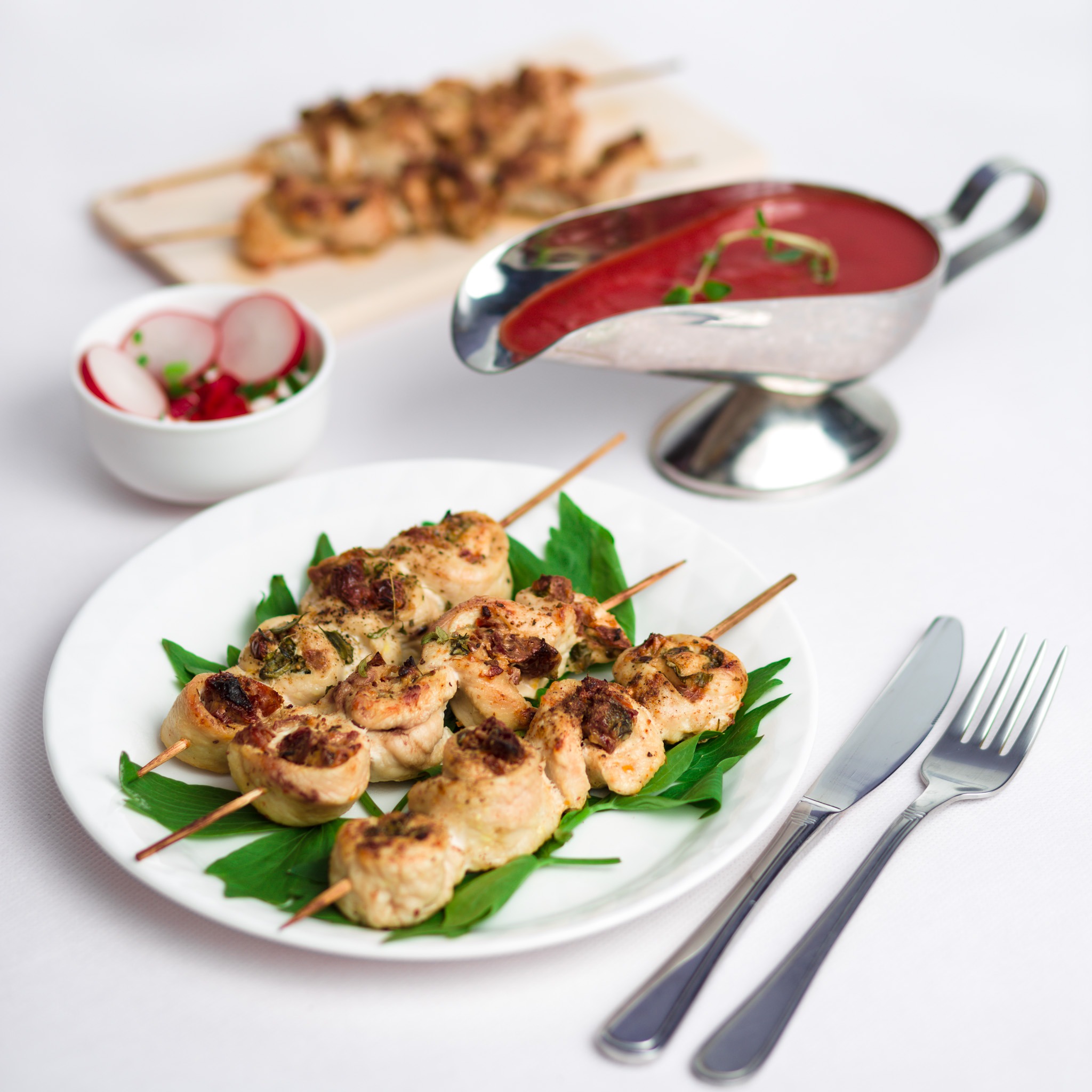 Elegancko podane szaszłyki z kurczaka na białym talerzu z zielonymi liśćmi, obok sos w sosjerce, rzodkiewki w miseczce i sztućce.