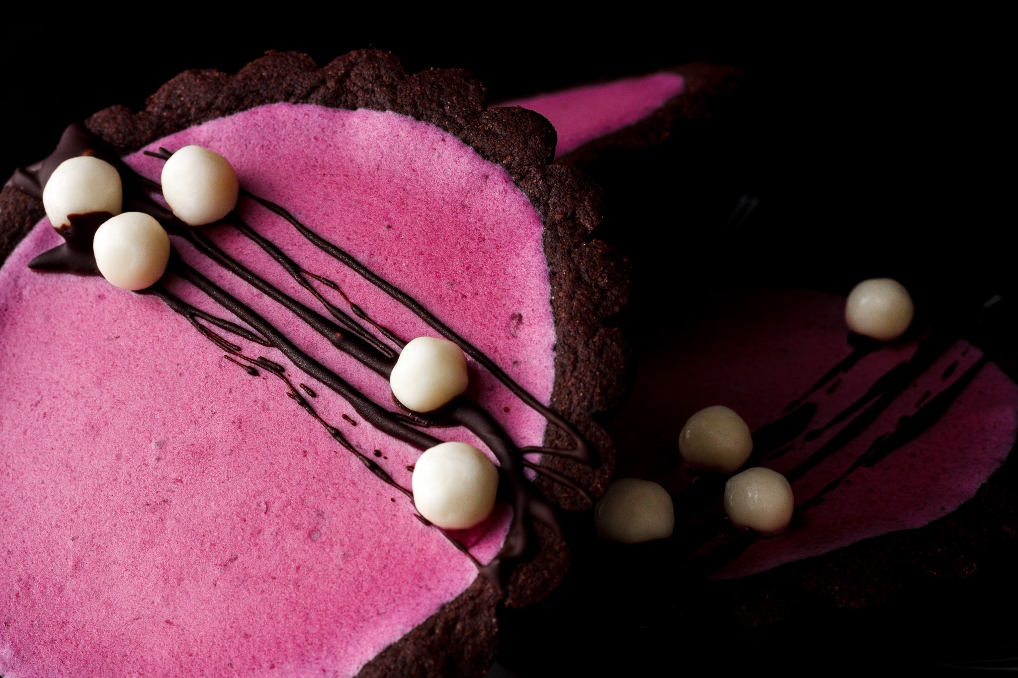 Ciemne, kruche ciasto z różowym kremem, polane czekoladą i udekorowane białymi kulkami, na czarnym tle.