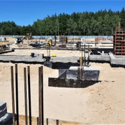 Fundamenty domu w trakcie budowy z widocznymi zbrojeniami, bloczkami betonowymi, deskowaniem i żółtą koparką w tle.