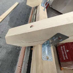 Drewniana konstrukcja dachu w trakcie montażu, widoczne elementy z drewna łączone metalowymi złączkami na śruby, w tle cegły i materiały budowlane Porotherm.