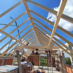 Konstrukcja drewniana dachu w trakcie budowy, widok od wewnątrz na więźbę dachową, murarzy pracujących na rusztowaniu i dźwig budowlany na tle błękitnego nieba.