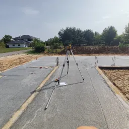 Świeżo wylana betonowa płyta fundamentowa domu jednorodzinnego z wystającymi prętami zbrojeniowymi, na której stoi niwelator laserowy na statywie, obok leżą notatki i miarka.