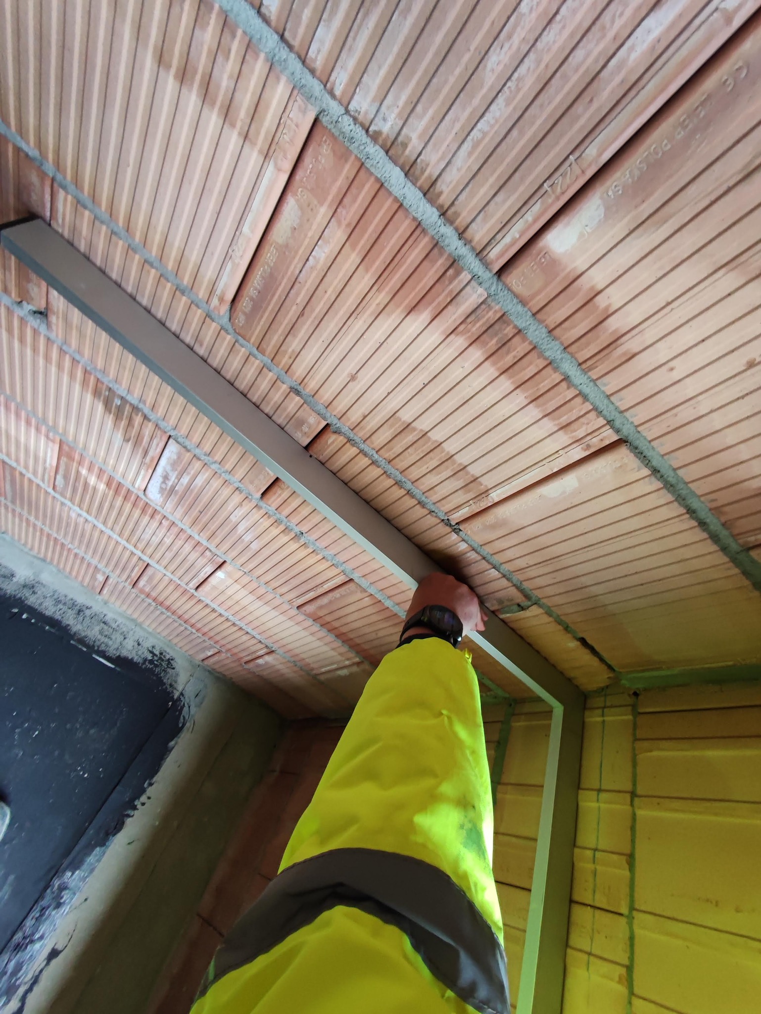 Montaż metalowego profilu pod sufitem z cegieł dziurawki, widoczna ręka pracownika w żółtej kurtce.