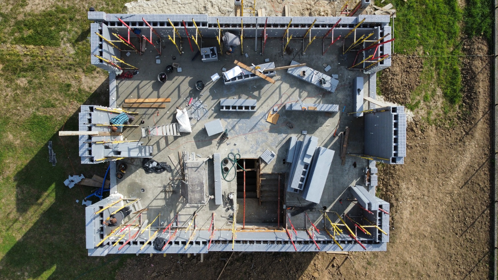Widok z góry na plac budowy domu jednorodzinnego z szalunkami traconymi, widoczne zbrojenia, narzędzia i materiały budowlane, dwóch pracowników w trakcie wykonywania prac.