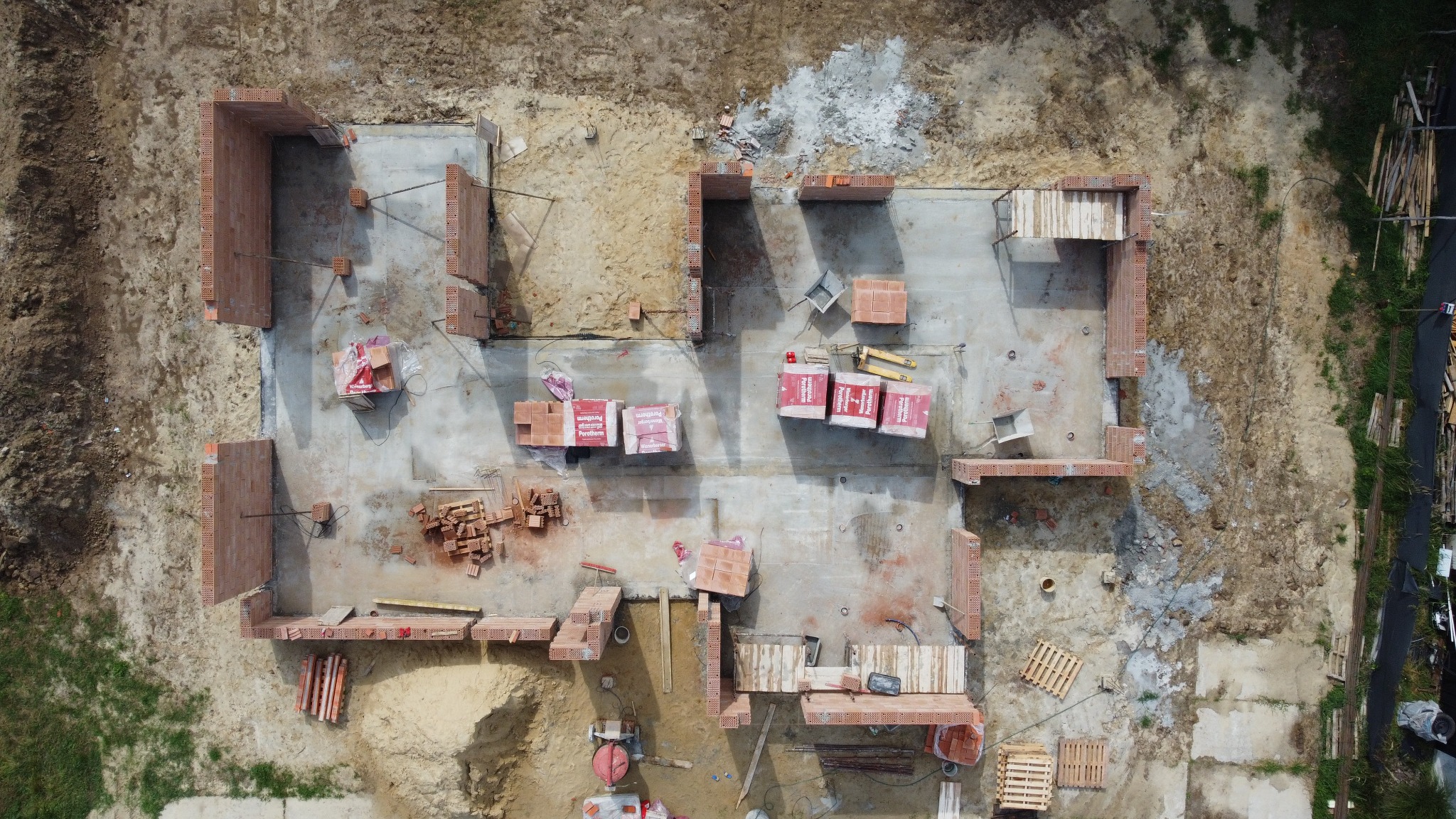 Wizualizacja z drona: budowa domu jednorodzinnego w trakcie wznoszenia ścian z czerwonej cegły Porotherm, widoczne palety z cegłami, zaprawa, narzędzia i fundamenty.