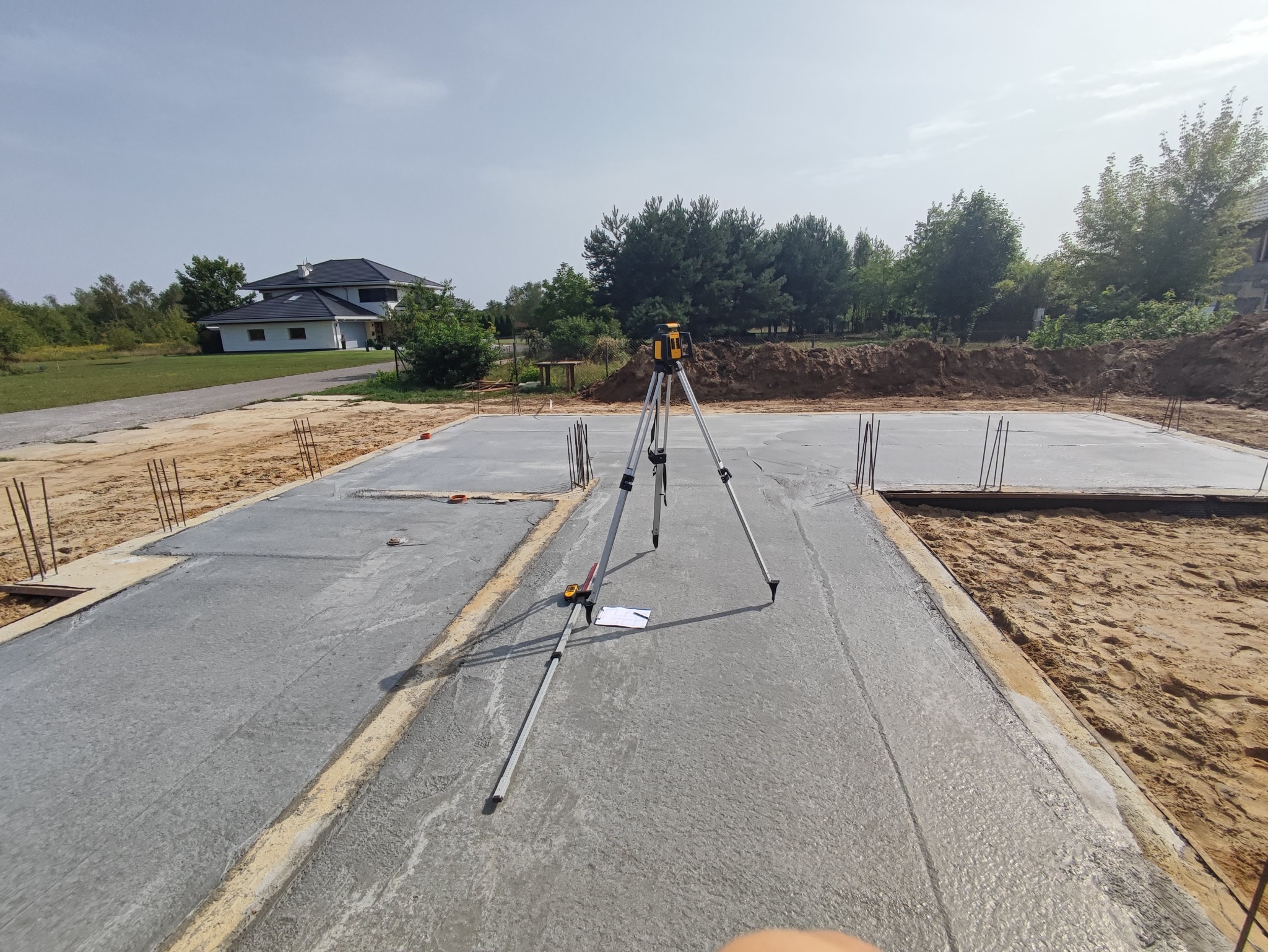 Świeżo wylana betonowa płyta fundamentowa domu jednorodzinnego z wystającymi prętami zbrojeniowymi, na której stoi niwelator laserowy na statywie, obok leżą notatki i miarka.