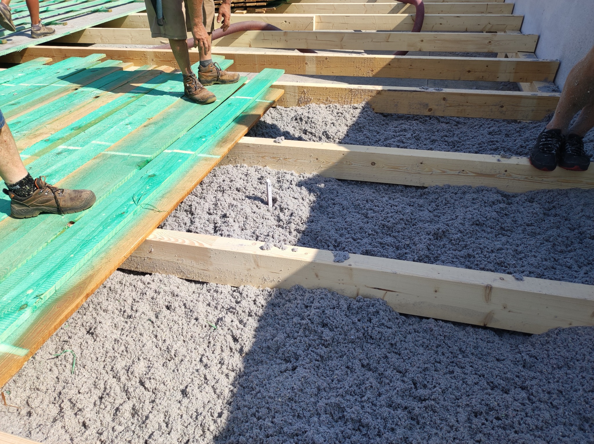 Montaż izolacji z wełny celulozowej na poddaszu, widoczne drewniane legary i fragment desek podłogowych pomalowanych na zielono.