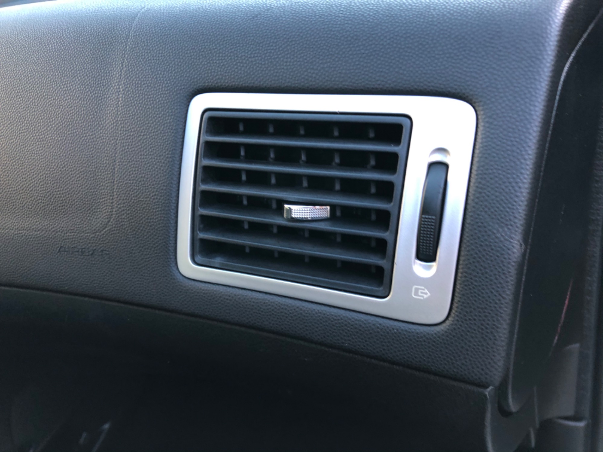 Kratka nawiewu powietrza w samochodzie z chromowaną ramką i regulacją, wkomponowana w czarną, teksturowaną deskę rozdzielczą.