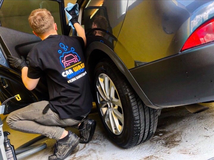 Pracownik w czarnej koszulce z logo myjni samochodowej czyści wnętrze czarnego auta szmatką, klęcząc obok otwartych drzwi.