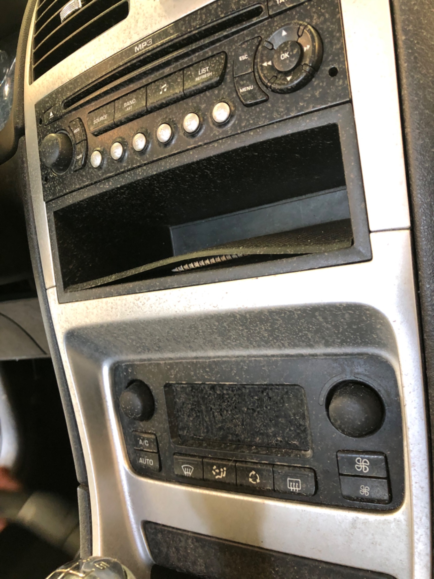 Detail deski rozdzielczej samochodu z widocznym radiem, panelem klimatyzacji i schowkiem, pokryte grubą warstwą kurzu.